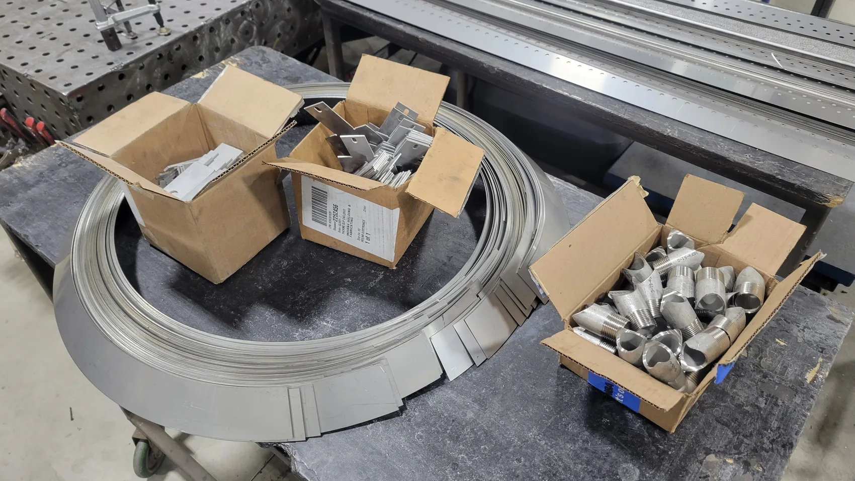 The Heat Wagon burner kit staged for assembly: rolled stainless strip, perforated strips, and threaded NPT nipples, cut and prepped in batches.