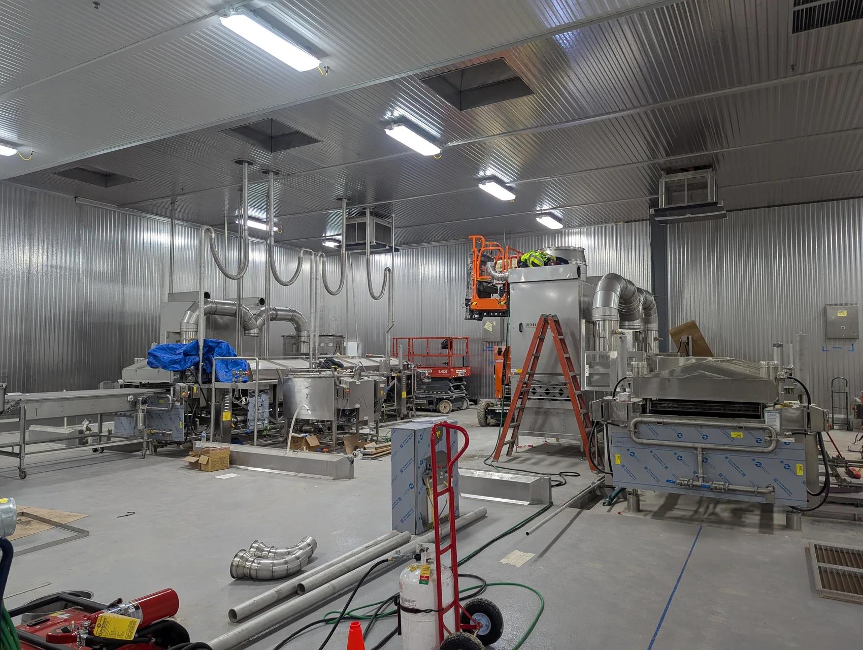 Wide view of a stainless cold-storage food plant with spiral stainless ductwork, fryer line and an orange boom lift during a field install by Sons of Thunder Welding