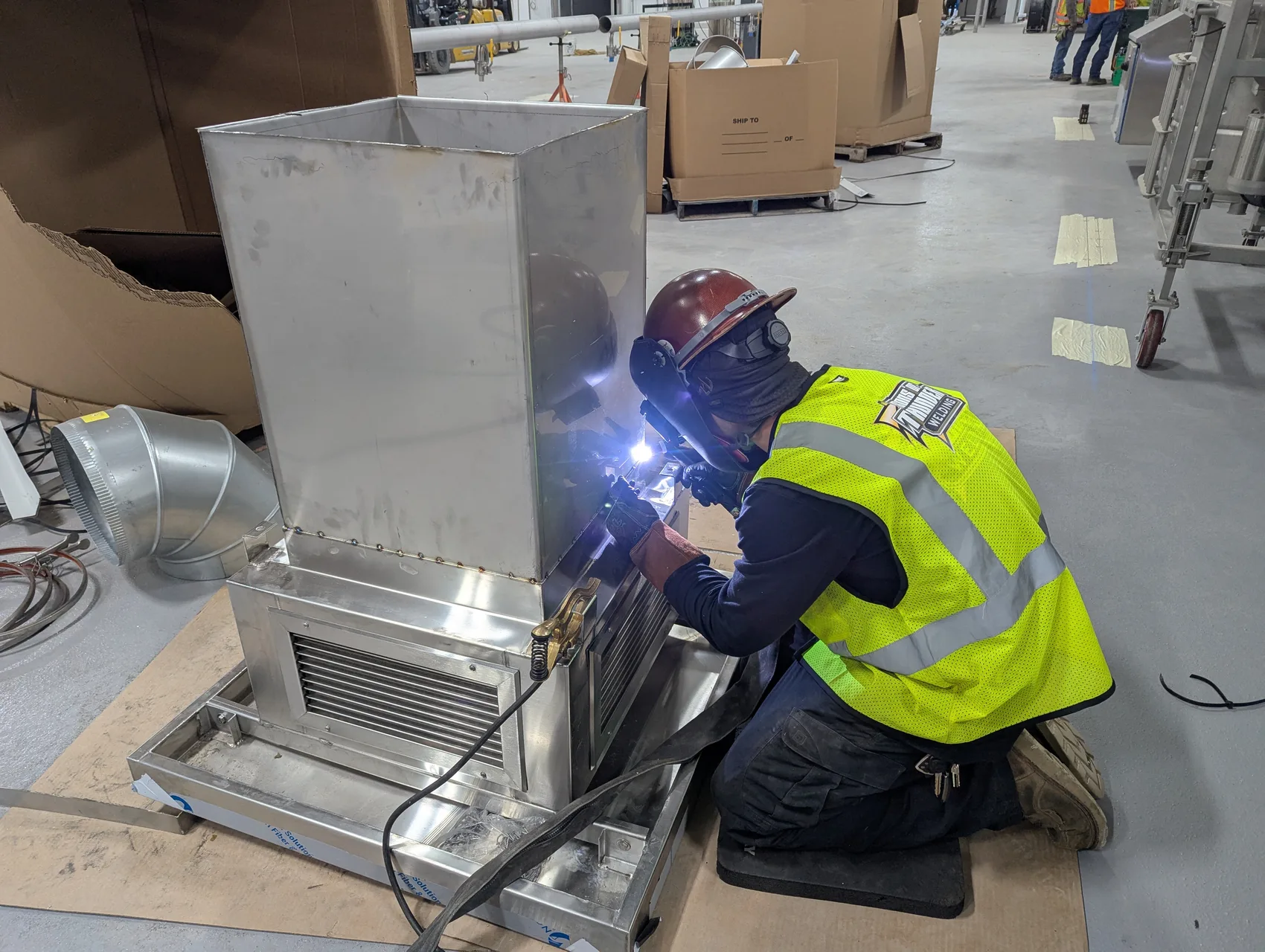 Welding the base of a stainless fryer hood, tying it into the round duct runs alongside.