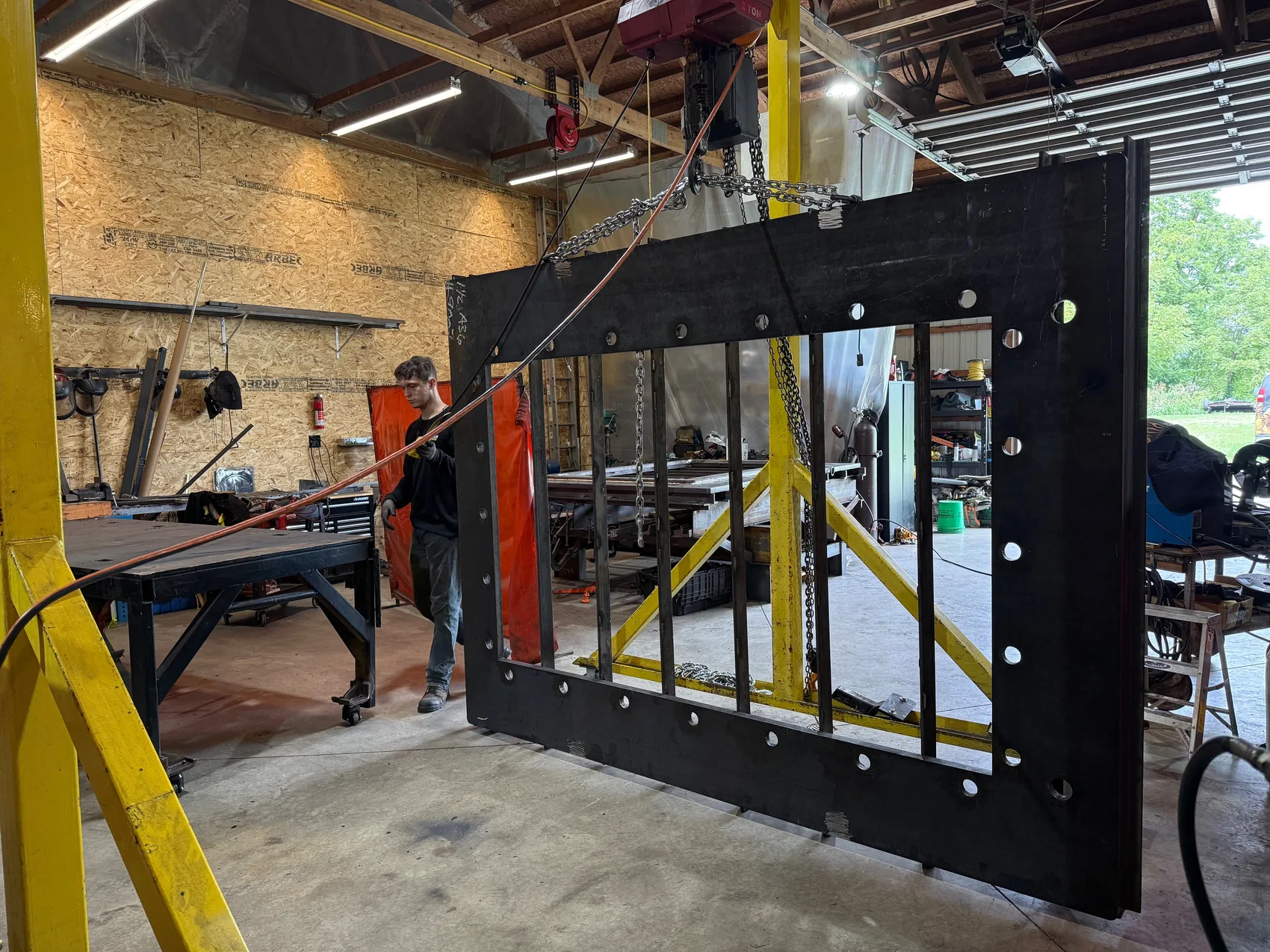 Large black steel rectangular roof-hatch reinforcement frame hanging from chain hoists in a welding shop while a worker guides it