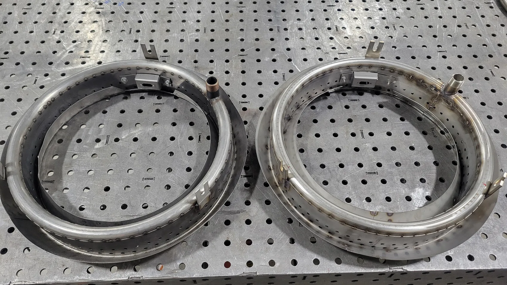 Two stainless steel helical burner assemblies with perforated bands on a shop fixture table by Sons of Thunder Welding